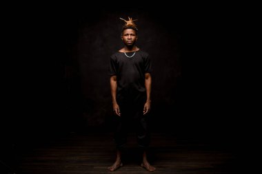 Siyah adam, sarı saçlı, siyah elbiseli, kameraya bakıyor.