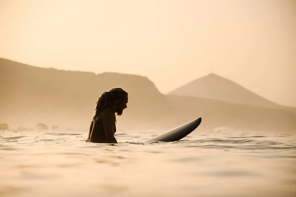 Vista lateral homem nu com dreadlocks sentado na prancha de surf na hora do pôr do sol — Fotografia de Stock