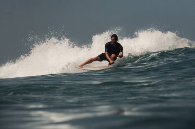 Siyah dalış kıyafetli, sakallı, sörf yaparken tek bacağı üzerinde çömelmiş bir adam.