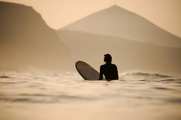 Vue arrière femme en combinaison de plongée noire assise sur la planche de surf au coucher du soleil — Photo