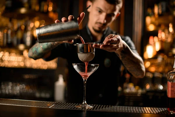 Чоловік бармен поливає алкогольний напій зі сталевого шейкера до склянки через сито — стокове фото