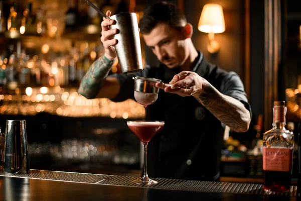 Barman tatuado derramando uma bebida alcoólica da coqueteleira de aço para o vidro através da peneira — Fotografia de Stock