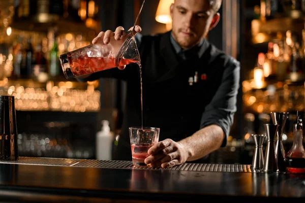 Cantinero masculino profesional que vierte una bebida alcohólica de la taza de medir al vaso a través del filtro colador —  Fotos de Stock