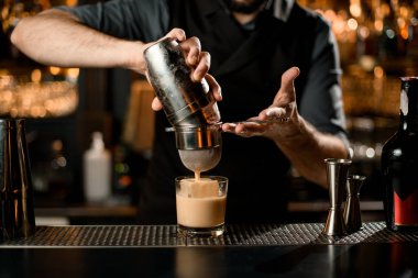 Profesyonel erkek barmen kremsi renkli alkolik bir içeceği süzgeç filtresinden geçirerek çelik karıştırıcıdan cama döküyor.