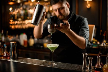 Erkek barmen kremsi yeşil renkli alkollü içeceği süzgeç filtresinden geçirerek bardağa döküyor.