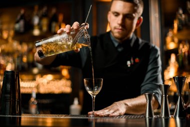 Profesyonel barmen sarı şeffaf alkollü içeceği süzgeç filtresinden ölçüm kabından cama döküyor.