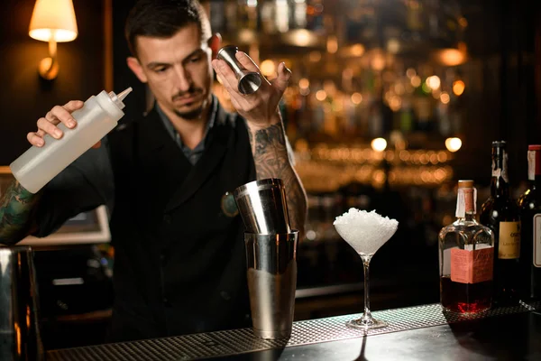 Barman masculino derramando uma bebida alcoólica do jigger para uma coqueteleira de aço segurando uma garrafa de plástico branco — Fotografia de Stock