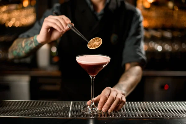 Professionele mannelijke barman versiert een heerlijke alcoholische cocktail in het glas met een gedroogde sinaasappel met pincet — Stockfoto