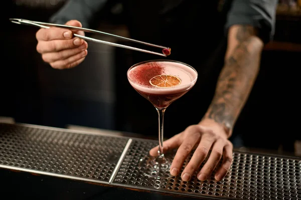 Barman decorando um coquetel alcoólico no copo com uma laranja seca e especiarias vermelhas com pinças com botão de rosa — Fotografia de Stock