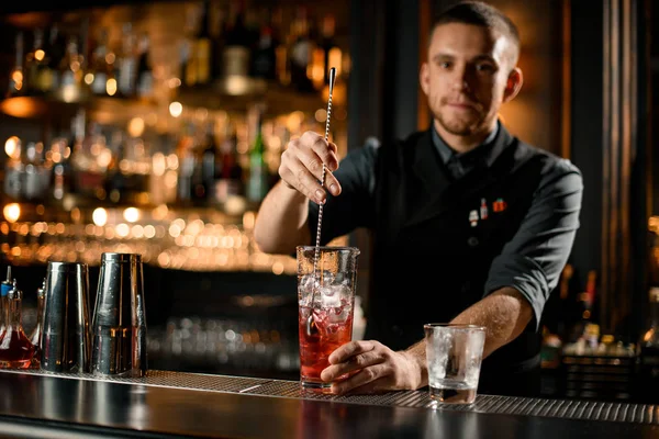 Cantinero profesional masculino revolviendo una bebida alcohólica roja en el vaso con la cuchara —  Fotos de Stock