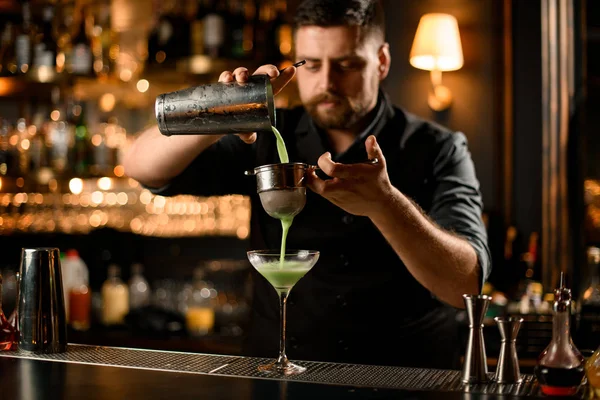 Barman professionnel versant une boisson alcoolisée de couleur vert crème de l'agitateur en acier au verre à travers le filtre à passoire — Photo