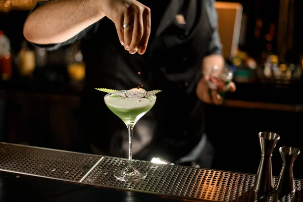 Barista che serve il cocktail di colore verde cremoso decorato con una foglia e arancia essiccata spruzzando su di esso con piselli al pepe rosso — Foto Stock