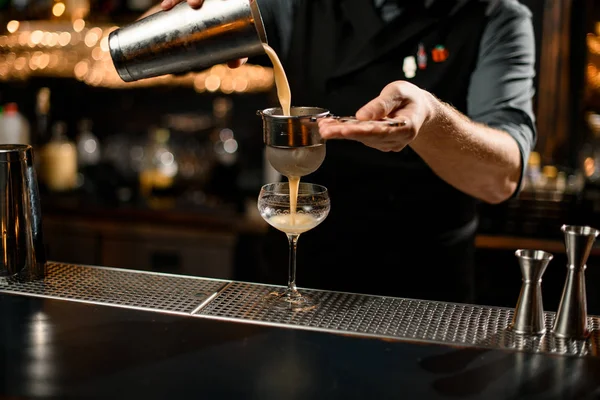 Επαγγελματίας μπάρμαν ρίχνει ένα κρεμώδες αλκοολούχο ποτό χρώμα από το shaker χάλυβα στο γυαλί μέσα από το φίλτρο σουρωτήρι — Φωτογραφία Αρχείου