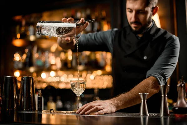 Professionele barman met een baard die alcoholische drank uit de maatbeker naar het glas giet door het zeeffilter — Stockfoto
