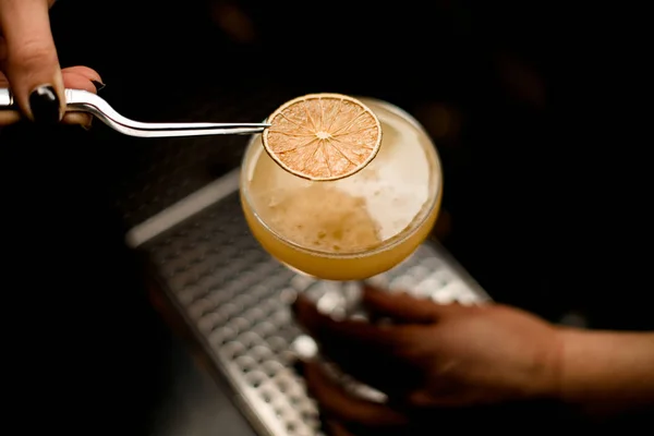 Ο bartender διακόσμησε το κίτρινο κρεμώδες αλκοολούχο κοκτέιλ με μια αποξηραμένη φέτα πορτοκαλιού με τσιμπιδάκι — Φωτογραφία Αρχείου