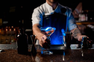 Profesyonel erkek barmen, mavi ışıkların altında uzun bardakta buz küpleriyle kahverengi alkollü kokteyl servis ediyor.