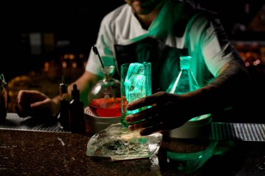 Profesyonel barmen, şeffaf bir alkollü içeceği yeşil ışıkta bardağa döküyor.