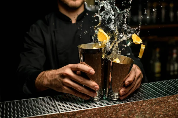 Homme barman tenant dans les mains un shakers en acier éclaboussant le cocktail avec des morceaux d'orange en elle — Photo