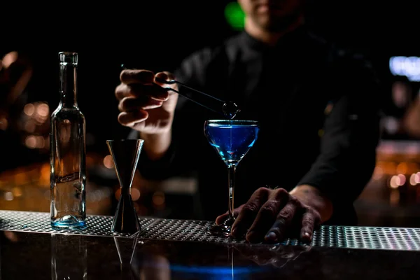 Cantinero lanzando a la bebida alcohólica azul una baya con pinzas —  Fotos de Stock