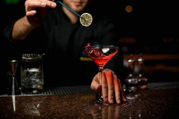 Professzionális férfi csapos szolgálja fel a vörös alkoholos koktélt jégkockákkal a magas pohárban citromszelet hozzáadása — Stock Fotó