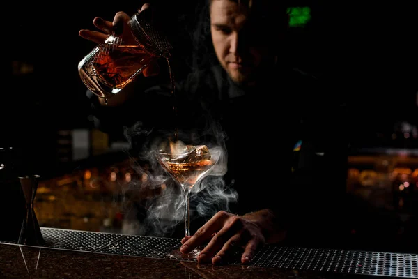 Професійний бармен наливає коричневий алкогольний напій з мірної чашки на склянку в парі — стокове фото