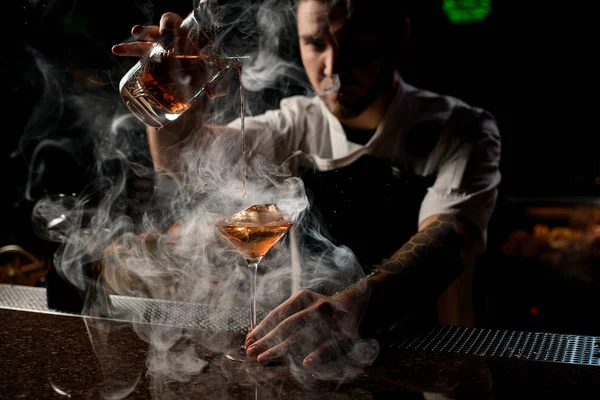 Cantinero masculino profesional que vierte un cóctel alcohólico marrón de la taza de medir al vaso en el vapor — Foto de Stock