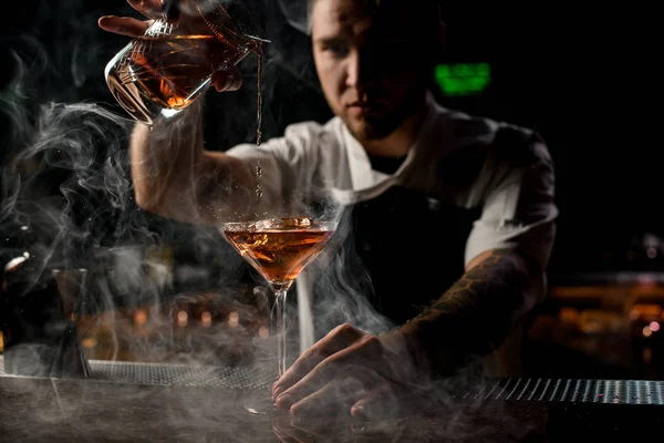Professzionális csapos, aki barna alkoholos koktélt tölt a mérőpohárból a gőzben lévő pohárba. — Stock Fotó