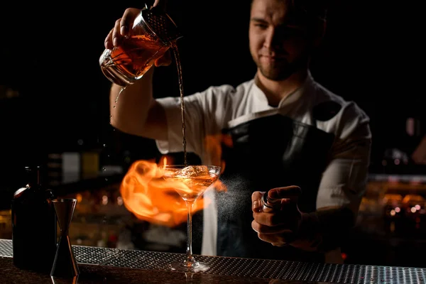 Profesionální barman nalévá hnědý alkoholický koktejl z odměrky na sklo, které hoří oheň — Stock fotografie