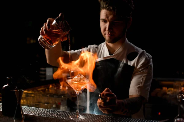 Barista professionista che versa un cocktail alcolico marrone dal misurino al bicchiere che brucia il fuoco — Foto Stock