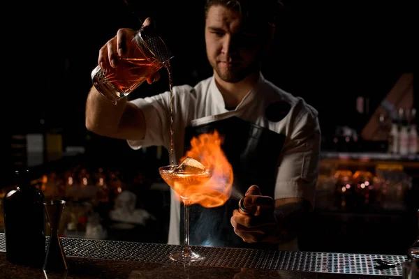 Cantinero macho vertiendo un cóctel alcohólico marrón desde la taza de medir hasta el vaso quemando el fuego — Foto de Stock