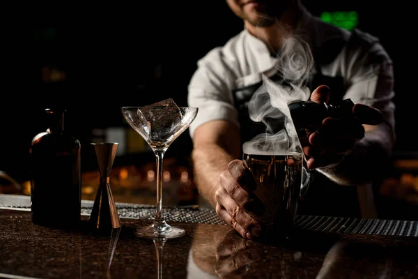 Male bartender holding in hands a steel shaker open it with the steam