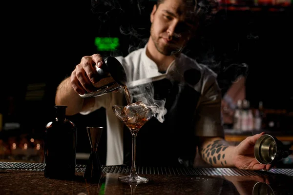 Cantinero macho que vierte una bebida alcohólica marrón del agitador de acero al vaso con el vapor —  Fotos de Stock