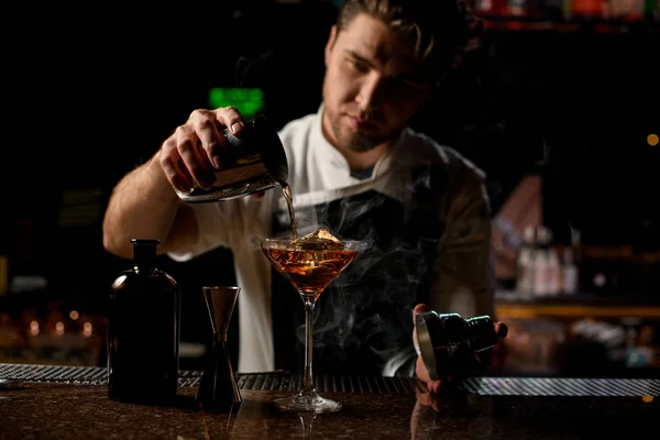 Professioneller männlicher Barkeeper, der ein braunes alkoholisches Getränk aus dem Stahlshaker in das Glas mit dem Dampf gießt — Stockfoto