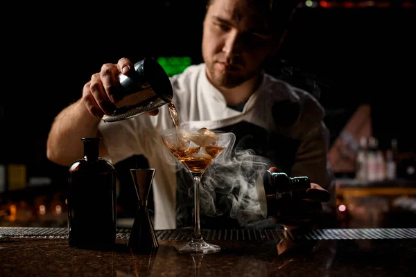 Професійний бармен наливає коричневий алкогольний напій зі сталевого шейкера на склянку з парою — стокове фото
