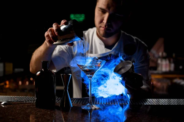 Cantinero profesional que vierte una bebida alcohólica marrón del agitador de acero al vaso bajo luz azul —  Fotos de Stock