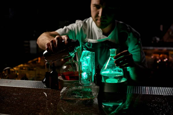 Cantinero masculino profesional que vierte una bebida alcohólica transparente del agitador de acero al vaso bajo luz verde —  Fotos de Stock