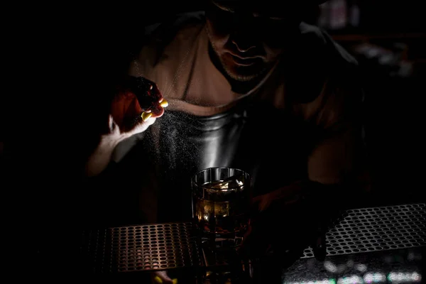 Cantinero rociando sobre la bebida alcohólica marrón con un jugo de ralladura de naranja en la oscuridad —  Fotos de Stock