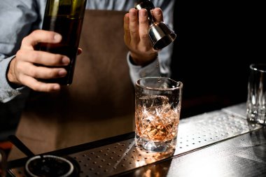 Profesyonel barmen, bir bardağa çelik jigger 'dan kahverengi alkollü bir içecek dolduruyor.