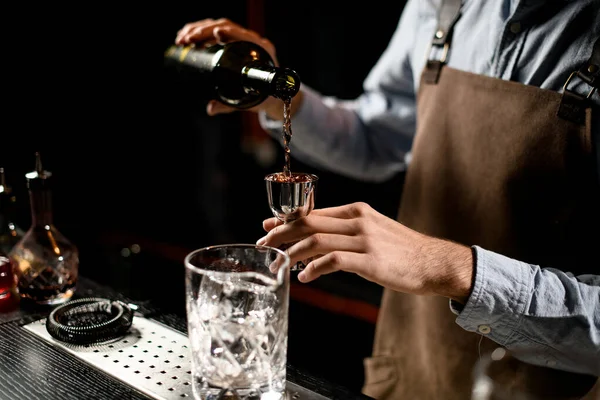 Cantinero masculino profesional sirviendo una bebida alcohólica marrón de la botella a un jigger de acero — Foto de Stock