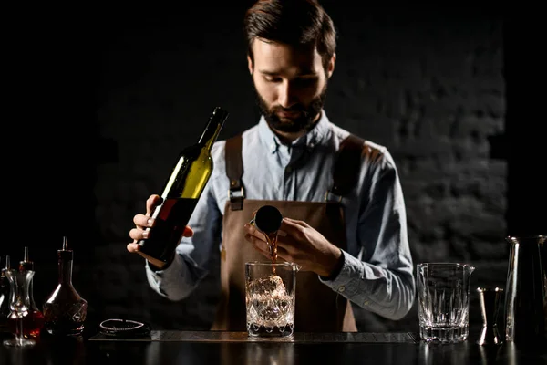 Barman masculino derramando uma bebida alcoólica dourada do jigger de aço para um copo de vidro — Fotografia de Stock