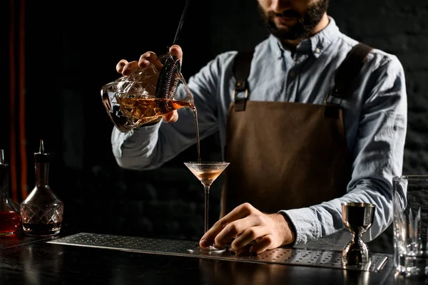 Cantinero masculino profesional sirviendo una bebida alcohólica marrón de la taza de medir con straner a una copa de martini —  Fotos de Stock