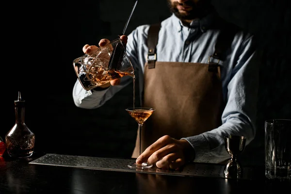 A csapos egy barna alkoholos italt tölt a mérőpohárból stranerrel egy martini pohárba. — Stock Fotó