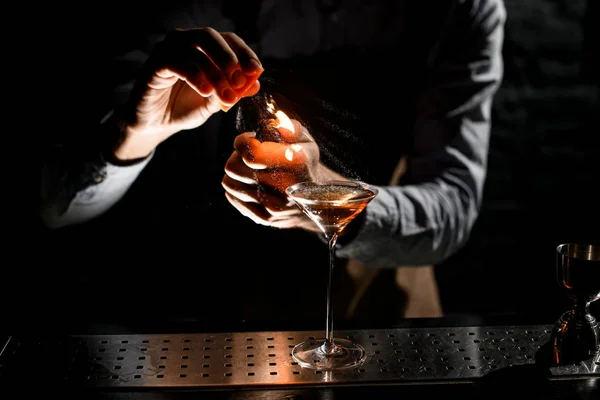 Barista maschio professionale che spruzza su un accendino con succo di scorza di orage a bevanda alcolica dorata in un bicchiere da martini — Foto Stock