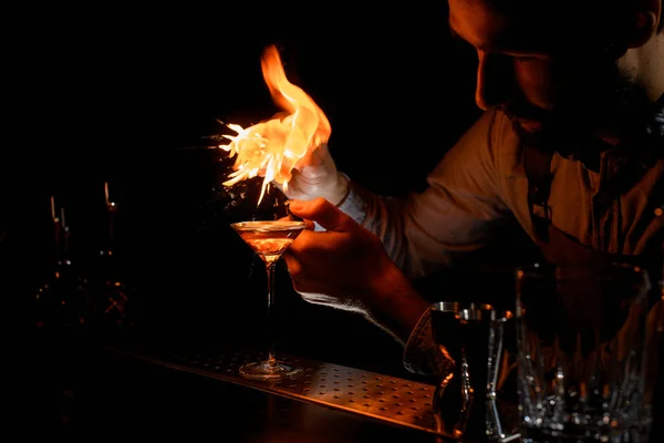 Male bartender spraying on a lighter with orage zest juice to the golden alcoholic drink in a martini glass — 스톡 사진