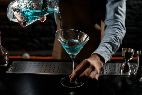 Cantinero masculino profesional sirviendo un cóctel alcohólico azul de la taza de medir con colador a una copa de martini — Foto de Stock