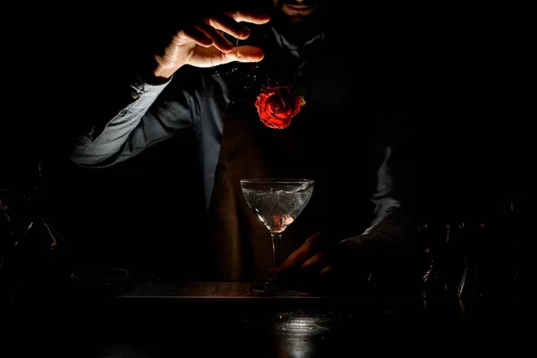 Cantinero masculino profesional lanzando un capullo de rosa roja a una copa de martini con un cóctel en la oscuridad —  Fotos de Stock