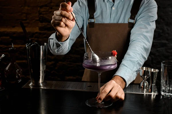 Barista maschio mescolando una bevanda alcolica viola nel bicchiere da cocktail grande decorato con un fiore rosso — Foto Stock