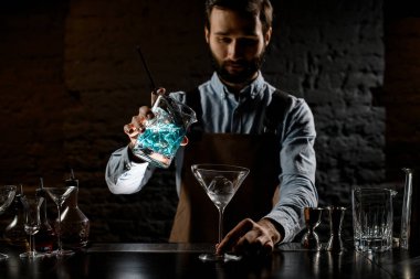 Profesyonel barmen, ölçü kabından martini bardağına mavi renkli alkollü kokteyl dolduruyor.