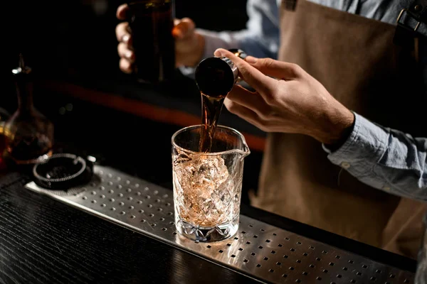 Cantinero profesional que vierte una bebida alcohólica marrón dorada del jigger de acero a un vaso — Foto de Stock
