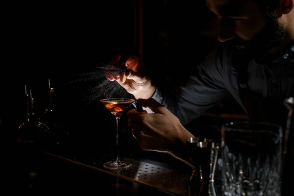Μπάρμαν ψεκασμό στο αλκοολούχο ποτό σε ένα ποτήρι μαρτίνι με χυμό ξύσμα orage στο σκοτάδι — Φωτογραφία Αρχείου
